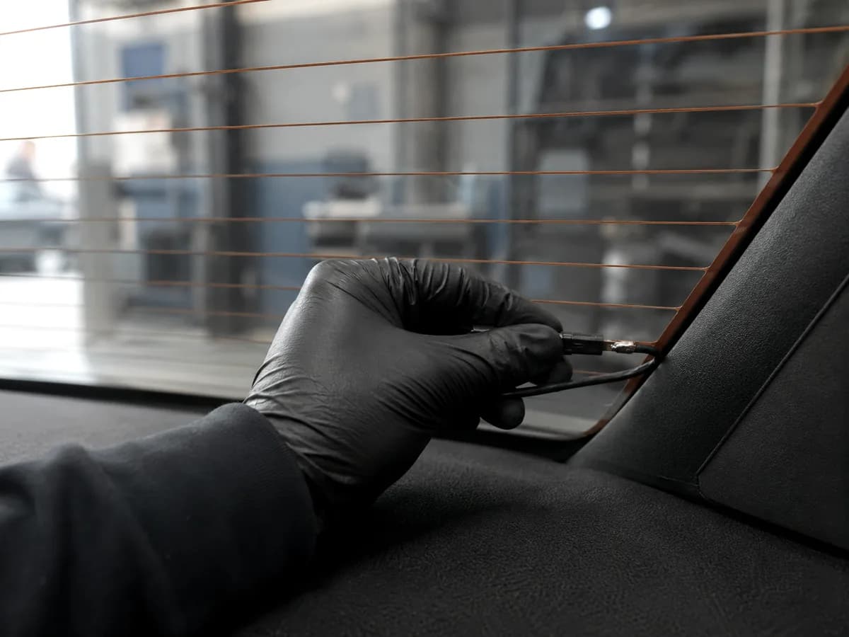A gloved hand working along the edge of a back glass, defroster lines running across it.